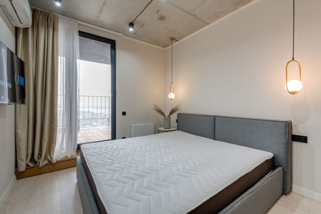 Bare mattress on a bed with a grey upholstered headboard beside a balcony window