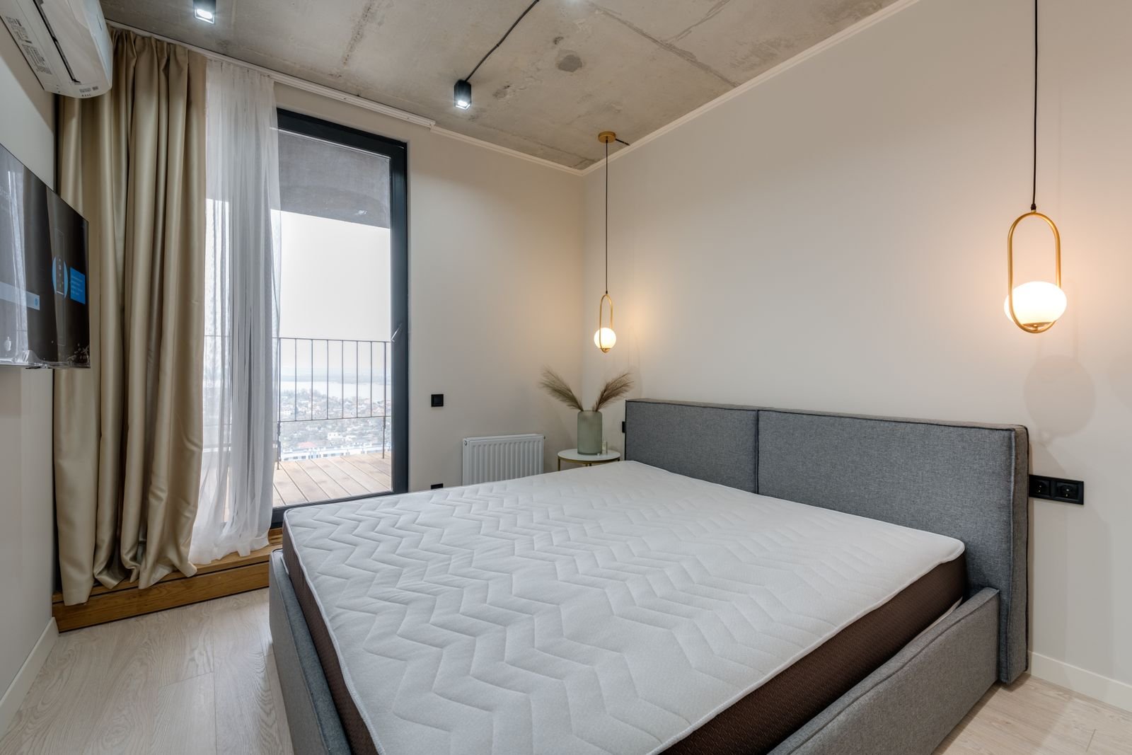 Bare mattress on a bed with a grey upholstered headboard beside a balcony window